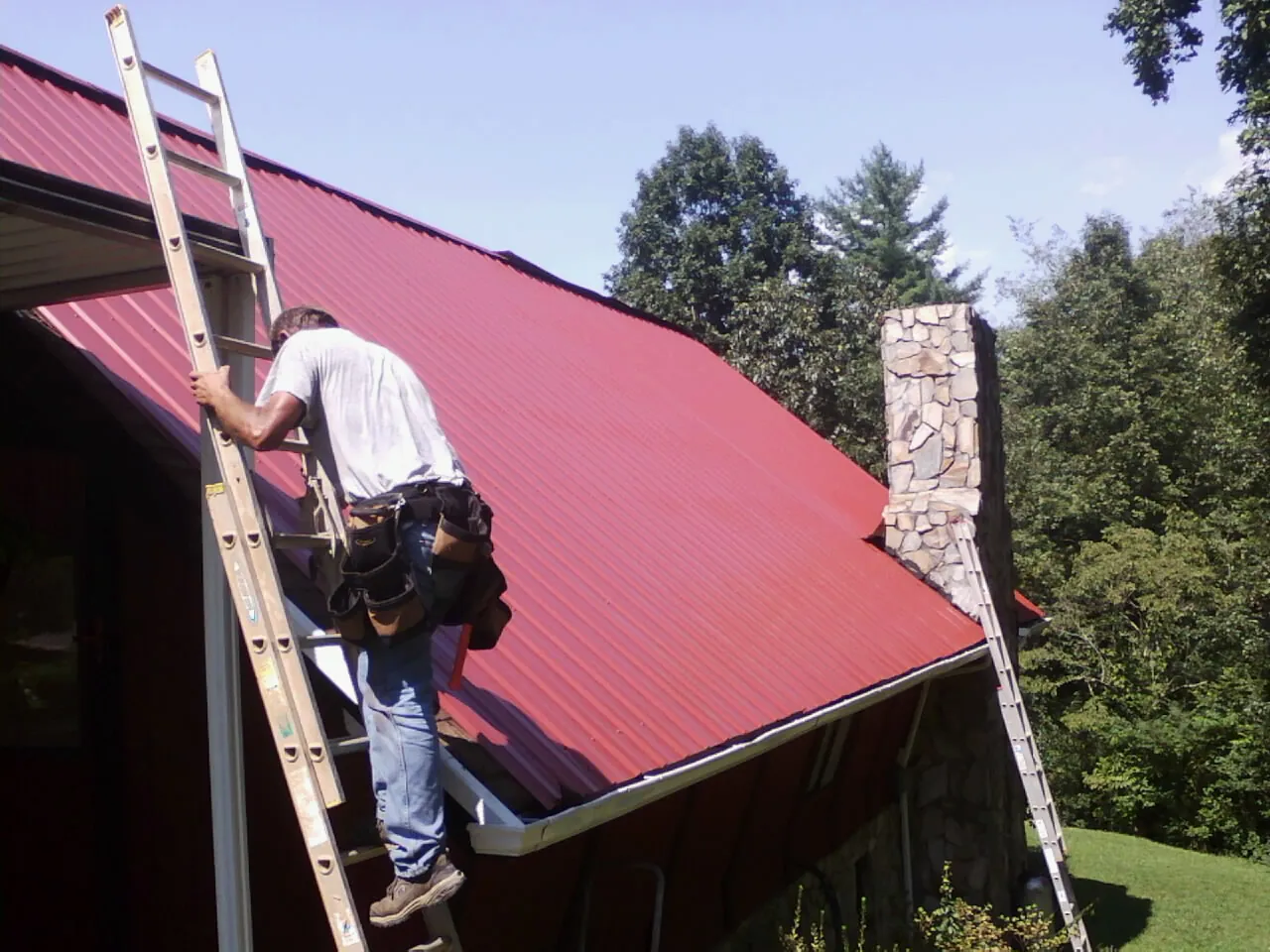 Roofer on a ladder beside a red metal roof and stone chimney — Soffit & Fascia Repair by Roofing Brothers in Boyds